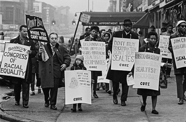 A black and white photograph of people from Brooklyn CORE walking with signs in a boycott against Sealtest Dairy Company in 1963 